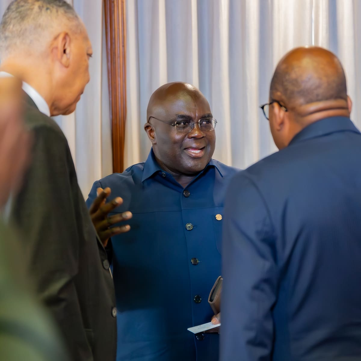 A smiling guest in glasses and a navy shirt, a business card in hand, talks with two delegates in dark suits seen from behind, in front of a pale curtain.