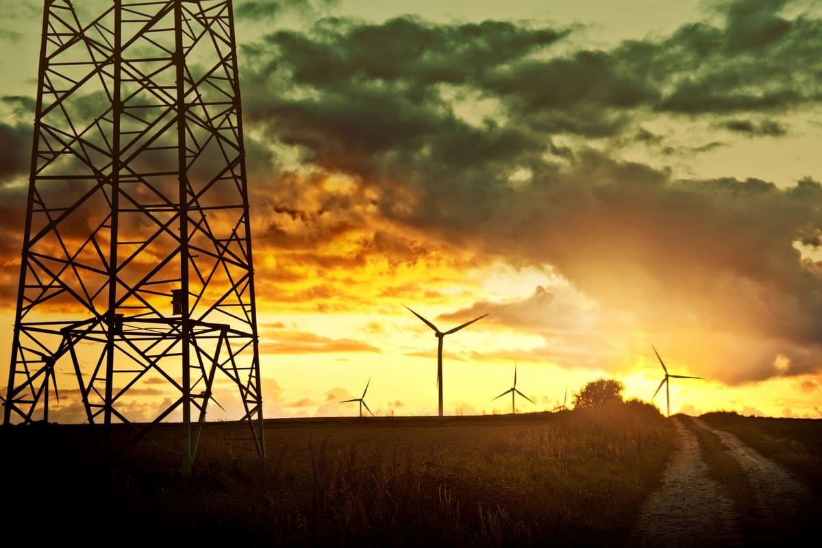 A high-voltage transmission tower and a line of wind turbines on the horizon at sunset