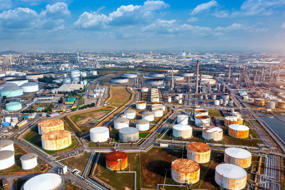 An aerial view of a fuel storage terminal, rows of round tanks stretching towards a city under a blue sky